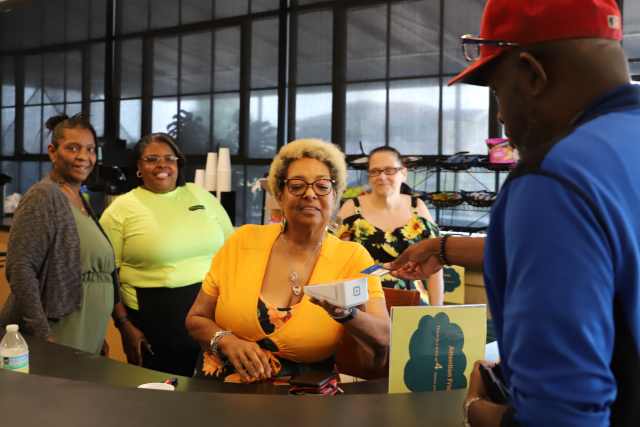 Three people smiling together at a laptop in a busy Clubhouse workspace
